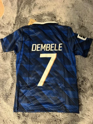Frankrike Dembele matchtröja blå - Säljer en blå och svart matchtröja från Nike med Dembele och nummer 7 på ryggen. Tröjan har vita detaljer, vit krage och guldiga tryck. Officiell fransk landslagströja med FFF-emblem och två stjärnor på bröstet. Perfekt för fotbollsfans! Passar M