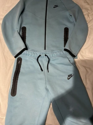 Ljusblått Nike loungewear-set - Säljer ett ljusblått loungewear-set från Nike med matchande ziptröja och byxor. Setet har svarta detaljer, dragkedjor på fickorna och Nike-logga på både tröja och byxa. Perfekt för chill dagar hemma eller när du vill vara bekväm och snygg. Nån fläck på tröjan vet inte om de går bort i tvätt annars är tröjan som ny. Byxorna är lite mer slitna ena snöret har gått sönder sen är de ett jätte litet hål vid knä området. Köpt från JD sport