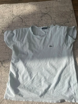 Ljusblå t-shirt från Lacoste - Säljer en ljusblå t-shirt från Lacoste med klassisk krokodillogo på bröstet. T-shirten har rund halsringning och korta ärmar. Perfekt för en clean och enkel stil.