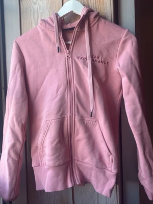 Rosa hoodie Peak Performance - Snygg rosa hoodie från Peak Performance med dragkedja framtill, broderad logga på bröstet och två fickor. Hoodien har huva med dragsko och ribbade muddar vid ärmslut och nederkant. Perfekt för en avslappnad och sportig stil.