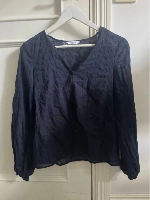 Mörkblå blus med broderade detaljer - Snygg mörkblå blus med v-ringning och diskreta broderade blommor i tyget. Blusen har långa ärmar och är lätt transparent, vilket ger en luftig känsla. Perfekt att matcha med jeans eller kjol för en trendig look.