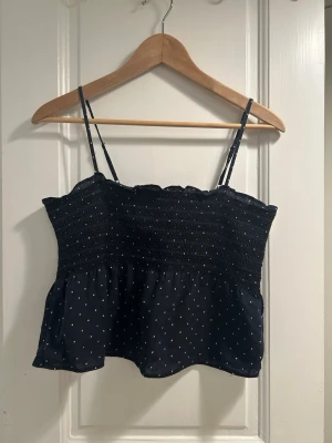 Svart smockad topp med prickar H&M - Säljer en svart smockad topp från H&M med tunna axelband och små vita prickar. Toppen har en stretchig överdel och ett lätt, flödande tyg nedtill. Perfekt för sommardagar och enkel att matcha med jeans eller kjol.