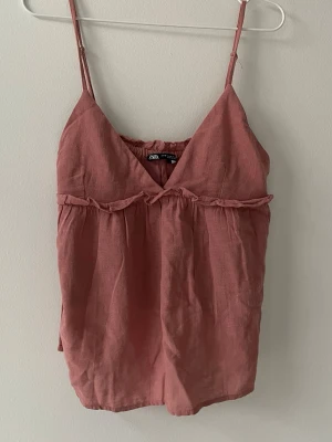 Rosa linnetopp från Zara - Söt rosa linnetopp från Zara med tunna axelband och v-ringning. Toppen har en volangdetalj under bysten och är tillverkad i ett luftigt linnematerial, perfekt för varma dagar. Enkel och trendig design som passar till mycket.