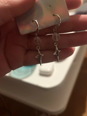 Silvriga stjärnörhängen med pärla - Säljer ett par snygga örhängen i silverfärg med hängande stjärnformad detalj och en genomskinlig pärla ovanför. Örhängena har en modern och trendig look, perfekta för att lyfta vilken outfit som helst. Materialet ser ut att vara metall och plast.