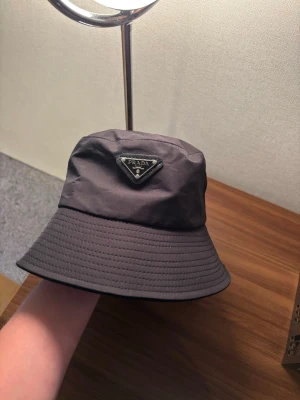 Svart bucket hat från Prada - Snygg svart bucket hat från Prada med det klassiska trekantiga logotyp-emblemet framtill. Hatten är tillverkad i ett glansigt, vattenavvisande material och har en bred, nedåtlutande brätte. Perfekt för en trendig och avslappnad stil.