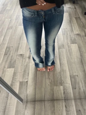 Blå bootcut jeans - Säljer ett par blå bootcut jeans med snygga slitningar och ljusa tvättade partier framtill och på sidorna. Jeansen har klassisk femficksmodell och låg midja. Perfekta för en avslappnad och trendig look.