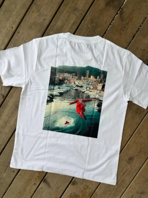 Charles leclerc f1 Vit Ferrari t-shirt med tryck - Snygg vit t-shirt med Ferrari-logga på bröstet och ett stort färgglatt tryck på ryggen som visar en person i röd overall vid en hamn. Klassisk passform och rund halsringning. Perfekt för dig som gillar streetwear och coola prints.