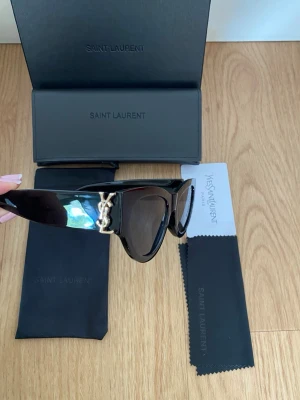 Svarta Saint Laurent solglasögon - Snygga svarta solglasögon från Saint Laurent med breda bågar och ikonisk YSL-detalj i guld på sidorna. Glasögonen har en klassisk, kantig form och kommer med originalfodral och putsduk. Perfekta för dig som vill ha en lyxig och trendig accessoar.
