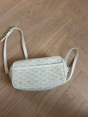 Vit axelväska med mönster - Snygg vit axelväska med justerbar rem och diskret mönster över hela väskan. Väskan har en rektangulär form och dragkedja framtill. Perfekt för dig som vill ha en stilren och trendig accessoar till din outfit.