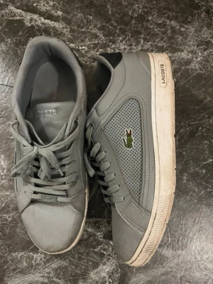 Gråa Lacoste sneakers med krokodil - Snygga grå sneakers från Lacoste med klassisk krokodillogo på sidan. Skorna har perforerade detaljer för extra ventilation, matchande grå snörning och vit sula med Lacoste-tryck. Perfekta för en clean och sportig stil.