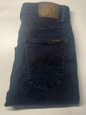 Nudie jeans  - Snygga mörkblå jeans från Nudie Jeans i slim fit. Klassisk femficksmodell med guldfärgade detaljer och Nudie-logga på bakfickan. Jeansen har hög midja och är tillverkade i stretchigt denim för en bekväm passform. Modellapp saknas. Midja 34cm, längd 99cm.  Ifall det inte finns någon bild i annonsen där jag bär plaggen jag säljer så är det för att jag ej kan ha storleken heller passformen, så kolla måtten för storlek och googla på modellnamnet för passform 😉 s3