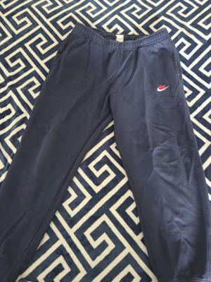 Mörkblå Nike mjukisbyxor - Mörkblå mjukisbyxor från Nike med elastisk midja och sidofickor. Byxorna har en klassisk passform och är prydda med Nike-logga i rött och vitt på vänster ben. Perfekta för en avslappnad stil och tillverkade i mjukt material.