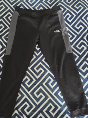Svarta mjukisbyxor The North Face - Svarta mjukisbyxor från The North Face med grå sidopaneler och dragkedjeficka på höger ben. Byxorna har en sportig look, resår i midjan och smal passform. Perfekta för chill eller träning.