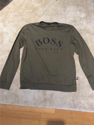 Olivgrön sweatshirt från Hugo Boss - Snygg olivgrön sweatshirt från Hugo Boss med stort svart BOSS-tryck på bröstet. Tröjan har rund halsringning, långa ärmar och ribbade muddar vid ärmslut och nederkant. Perfekt för en avslappnad och trendig look. Sitter M
