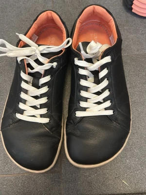 Groundies Barefoot Skor - Svarta sneakers med vit sula och vita skosnören. Skorna har en låg modell och insida i ljus orange färg. Tillverkade i slätt material med enkel och stilren design. Perfekta för en avslappnad och trendig look. Barefoot skor och bra kvalitet 