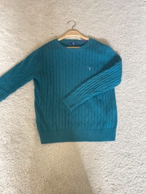 Grönblå kabelstickad tröja GANT - Snygg kabelstickad tröja från GANT i en djup grönblå färg. Tröjan har rund halsringning, ribbade muddar och diskret GANT-logga broderad på bröstet. Perfekt för lager på lager och ger en klassisk look.