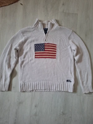 Vit stickad tröja Polo Ralph Lauren med Usa flagga i storlek Xs - Vit stickad tröja från Polo Ralph Lauren i 100% bomull. Tröjan har halv dragkedja, amerikansk flagga broderad på bröstet och RL-broderi vid nederkanten. Klassisk och avslappnad passform med ribbade muddar och krage. Tröjan är i storlek XS