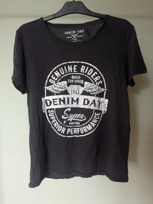 Svart t-shirt med tryck från Denim Day - Cool svart t-shirt från Denim Day med stort vitt tryck på bröstet. Trycket har texten 'Genuine Riders', 'Denim Day', 'Superior Performance' och detaljer med vingar. Klassisk rund hals och korta ärmar. Perfekt för en avslappnad streetstyle.