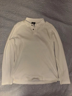 Vit långärmad piké från H&M - Klassisk vit långärmad pikétröja från H&M i slim fit-modell. Tröjan har krage, knappar vid halsen och är tillverkad i ett strukturerat bomullstyg som ger en snygg och stilren look. Perfekt för dig som gillar enkel och clean stil.