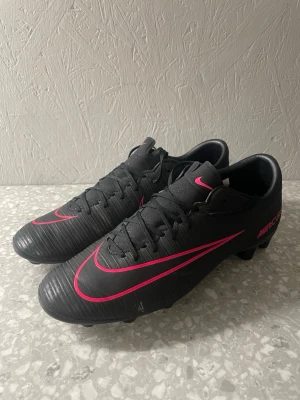 Svarta Nike Mercurial fotbollsskor - Säljer ett par svarta Nike Mercurial fotbollsskor med rosa detaljer och klassisk låg profil. Skorna har snörning, räfflad ovansida för bättre bollkontroll och dobbar för spel på gräs. Perfekta för dig som vill ha stilrena och snabba skor på planen.