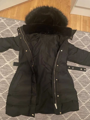 Svart lång dunjacka med svart päls - Snygg svart lång dunjacka med stor huva och fuskpäls. Jackan har dragkedja, knappar och bälte i midjan för en schysst passform. Perfekt för kalla vinterdagar och riktigt varm tack vare dunfyllningen och den höga kragen. Pris kan diskuteras!