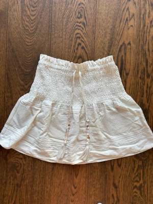 Vit kortkjol med knyt från Zara - Supersöt vit kortkjol från Zara med smock i midjan och dekorativa knytband med pärlor. Kjolen har volangkant och är gjord i ett lätt material som passar perfekt till varma dagar.