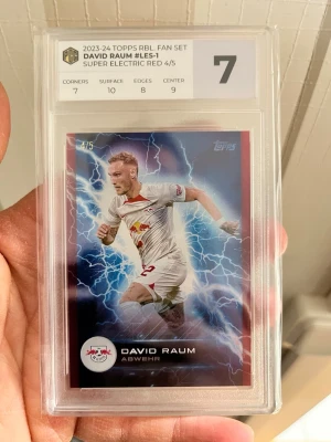 Topps David Raum Super Electric Red - Samlarkort från 2023-24 Topps RBL Fan Set med David Raum, Super Electric Red 4/5. Kortet är graderat av Professional Card Grading och har en röd ram med blixtmotiv. Perfekt för fotbollsfans och samlare.