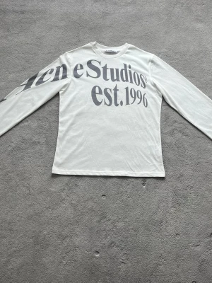 Vit långärmad tröja Acne Studios - Vit långärmad topp från Acne Studios med stor svart logotyp och text 'est. 1996' tryckt över bröstet och ena ärmen. Tröjan har rund halsringning och är gjord i mjuk bomull, perfekt för en clean och stilren look.