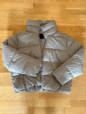 Beige pufferjacka från 157 - Säljer en beige pufferjacka från 157 med hög krage och dragkedja framtill. Jackan har en boxig passform och breda kanaler som ger en riktigt fluffig look. Perfekt för kalla vinterdagar och enkel att matcha med olika outfits.