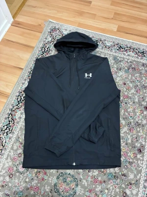 Svart hoodie från Under Armour - Säljer en svart hoodie från Under Armour med dragkedja framtill och justerbar huva. Hoodien har ett vitt logotryck på bröstet och två fickor på sidorna. Tillverkad i ett mjukt och bekvämt material, perfekt för chill dagar eller träning.