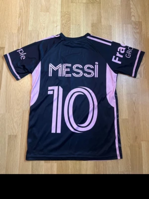 Svart Inter Miami Messi matchtröja - Säljer en svart och rosa matchtröja från Inter Miami med Messi och nummer 10 på ryggen. Tröjan har korta ärmar, rund hals och tryckta detaljer i rosa, inklusive klubbmärke och sponsorer. Tillverkad i ett lätt och ventilerande material, perfekt för fotboll eller träning.