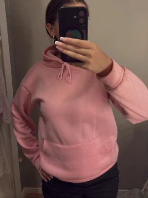 Rosa hoodie med huva - Säljer en rosa hoodie med klassisk känguruficka och dragsko i huvan. Hoodien har ribbade muddar vid ärmslut och nederkant. Perfekt för dig som gillar en enkel och clean stil. Materialet känns mjukt och bekvämt.