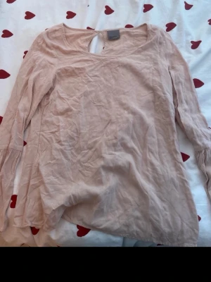 Ljusrosa blus från Vero Moda - Ljusrosa blus från Vero Moda med långärmad design och rund halsringning. Blusen har en liten knappdetalj i nacken och är tillverkad i ett tunt, mjukt tyg som ger ett avslappnat intryck. Perfekt att styla med jeans eller kjol.