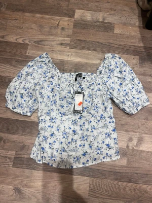 Blommig puffärm blus i vitt och blått - Supersöt blus med puffiga korta ärmar och smockad överdel. Vit botten med blått blommönster och små gröna detaljer. Fyrkantig ringning och lätt figursydd passform. Perfekt för en romantisk look och till jeans eller kjol.