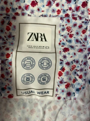 Blommig skjorta från Zara - Säljer en vit skjorta från Zara med små blommor i rött och blått. Skjortan har knappar framtill och klassisk krage. Perfekt för dig som gillar färgglada och lekfulla mönster. Tillverkad i bomull för en skön känsla.