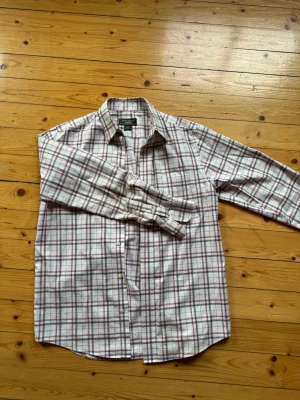 Rutig skjorta från Eddie Bauer - Klassisk långärmad skjorta från Eddie Bauer i vitt med rött och grått rutmönster. Skjortan har knappar framtill och är tillverkad i mjuk bomull. Perfekt för en avslappnad och stilren look.