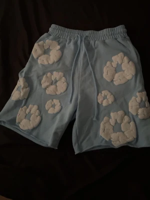 Ljusblå Denim Tears shorts med vita moln  - Unika ljusblå shorts med elastisk midja och snörning. Dekorerade med stora vita molnliknande applikationer över hela plagget för en lekfull vibe. Perfekta för sommarens alla äventyr och riktigt sköna att bära.