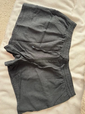 svarta linneshorts med snörning - Snygga svarta shorts i linne med elastisk midja och snörning framtill. De har en avslappnad passform och är perfekta för varma dagar. Shortsen har fickor på sidorna och är lätta och luftiga.