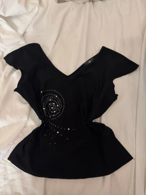 Vintage svart topp med v-ringning och stjärnmönster - Svart topp med djup v-ringning både fram och bak. Toppen har ett coolt spiral- och stjärnmönster i silverfärgade detaljer på framsidan. Kortärmad och figurnära passform, perfekt för att lyfta en outfit.