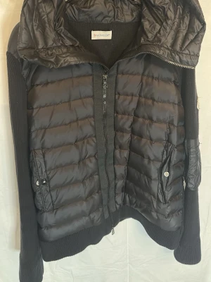 Svart pufferjacka från Moncler - Snygg svart pufferjacka från Moncler med stickade ärmar och sidor. Jackan har en stor huva, två fickor med knappar och dragkedja framtill. Perfekt för kalla vinterdagar och ger en sportig men stilren look.