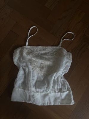 Vit linnetopp med spetsdetaljer - Söt vit linnetopp med tunna axelband och fina spetsdetaljer framtill. Toppen har en avslappnad passform och bredare kant nedtill. Perfekt att styla till jeans eller kjol för en fräsch look.