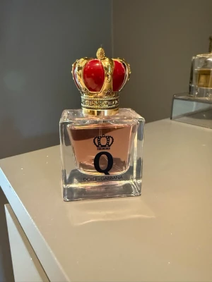 Dolce & Gabbana Q EdP 30ml - Lyxig parfym från Dolce & Gabbana i en fyrkantig glasflaska med ljusrosa doftvätska. Flaskan har en unik kork formad som en kungakrona i rött och guld, vilket ger en exklusiv känsla. Innehåller 30 ml Eau de Parfum. Förvarats svalt och mörkt.