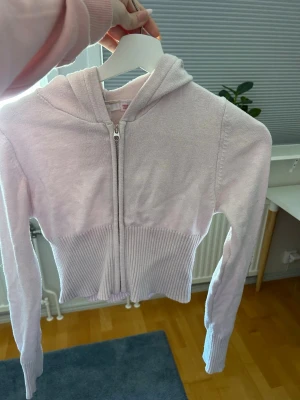Ljusrosa hoodie från Gina Tricot - Supersöt ljusrosa hoodie från Gina Tricot med dragkedja framtill och ribbade muddar i midjan och vid ärmsluten. Hoodien har en klassisk huva och är gjord i mjukt stickat material som känns skönt mot huden. Perfekt för en chill och avslappnad stil.