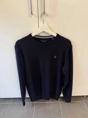 Mörkblå stickad tröja från Gant i Lam Ull, Stl M - Mörkblå stickad tröja från Gant i superfin lammull. Klassisk rund halsringning med ribbade muddar och diskret Gant-logga broderad på bröstet. Perfekt för dig som gillar stilrena och tidlösa plagg.