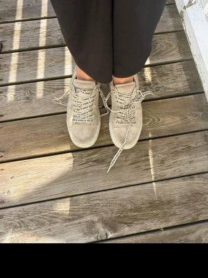Beige sneakers med textdetaljer - Säljer ett par beige sneakers med coola textdetaljer på skosnörena. Skorna har en klassisk låg modell och är tillverkade i mockaliknande material. Perfekta för dig som vill ha en unik touch på din outfit.