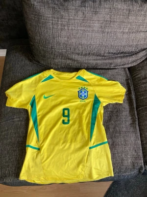 Brasilien Ronaldo nazario 9 - Gul Brasilien fotbollströja från Nike med gröna detaljer, nummer 9 och Ronaldo på ryggen. Tröjan har korta ärmar, rund hals och Brasilien-emblem på bröstet. Perfekt för dig som älskar fotboll och vill hylla en legend.
