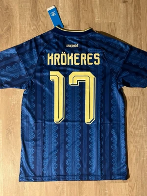 Blå Sverige fotbollströja Krökeres - Säljer en blå Sverige fotbollströja med gult tryck, nummer 17 och namnet KRÖKERES på ryggen. Tröjan har gul krage, tre gula ränder på axlarna och Sveriges landslagsemblem på bröstet. Tillverkad i ett lätt och ventilerande material.