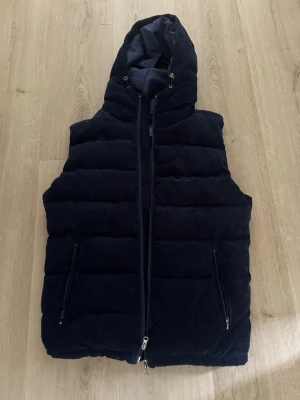 Mörkblå quiltad dunväst med huva - Snygg mörkblå dunväst. Västen är i princip som ny. Ny kostar 800 kr. Västen har dragkedja framtill och två fickor med dragkedja. Perfekt för lager på lager och ger en cool look till din outfit.