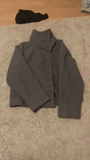 Grå ullkappa med hög krage - Stilren grå kappa i ullmix med hög krage och stora knappar framtill. Kortare modell med breda ärmslut och minimalistisk design. Perfekt för dig som gillar en clean och modern look.