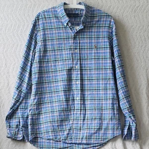 Blårutig skjorta från Ralph Lauren - Klassisk blårutig skjorta från Ralph Lauren med knappar framtill och button-down krage. Skjortan har långa ärmar och en liten broderad logga på bröstet. Tillverkad i mjuk bomull och har en avslappnad passform.